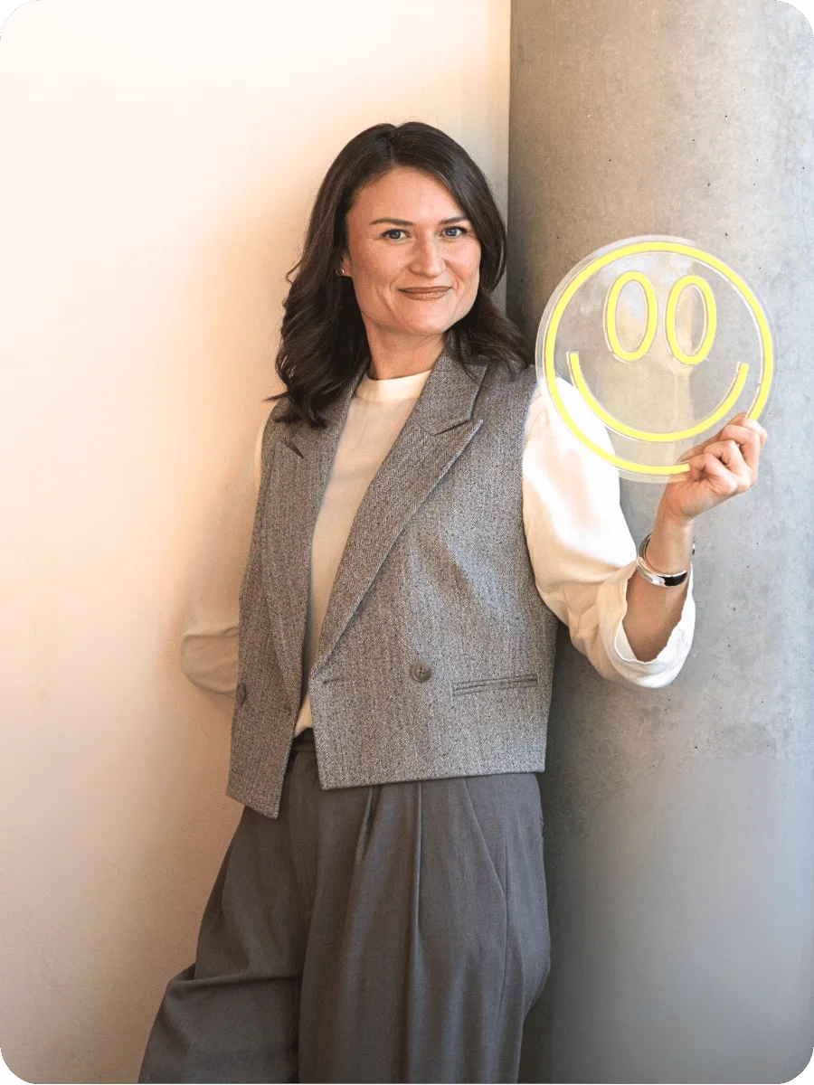 Juliana Bernhardt in grauer Weste lehnt an einer Betonsäule und hält ein Neon-Smiley